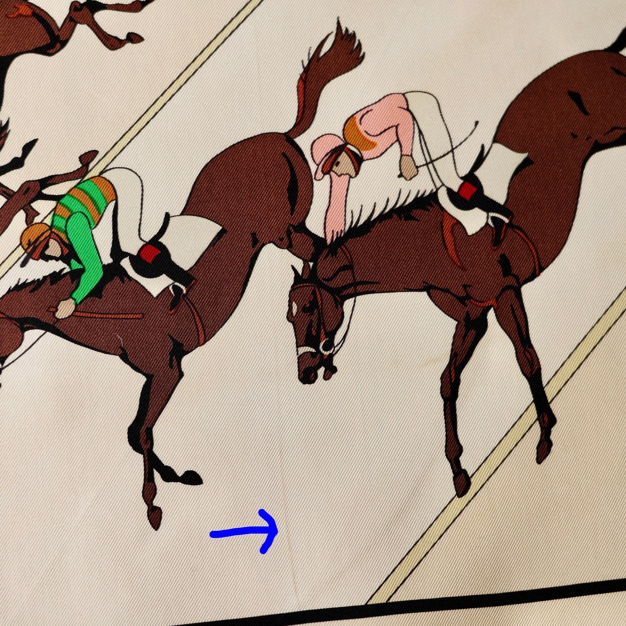 Zoom sur trace discrète sur la pliure sur le fond crème du foulard Hermès "Les Courses", pointée par deux flèches bleues.